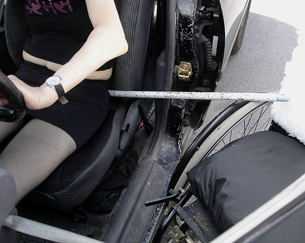 Användaren sitter på passagerarsätet, ett borstskaft som används som ryggstöd vid förflyttning ligger bakom henne och på rullstolen hjul