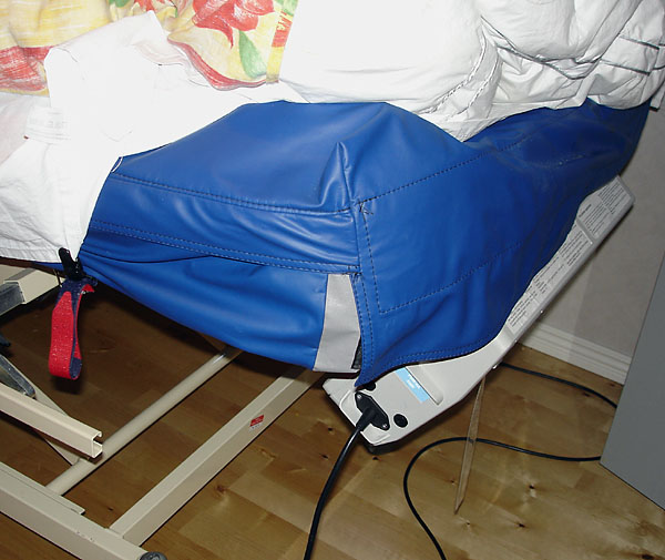 En tjock madrass med plastskydd ligger på en elektrisk reglerbar säng. En kompressor hänger på sängens fotända.