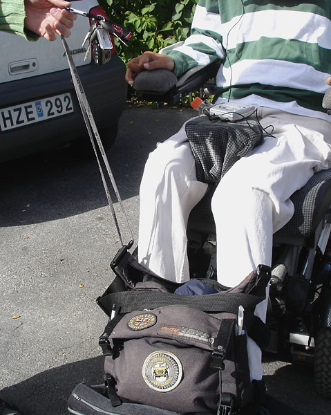 User seated in wheelchair. A person holds up a bunch of keys on a string fastened a bag. The bag is attached to the footplates of the wheelchair.