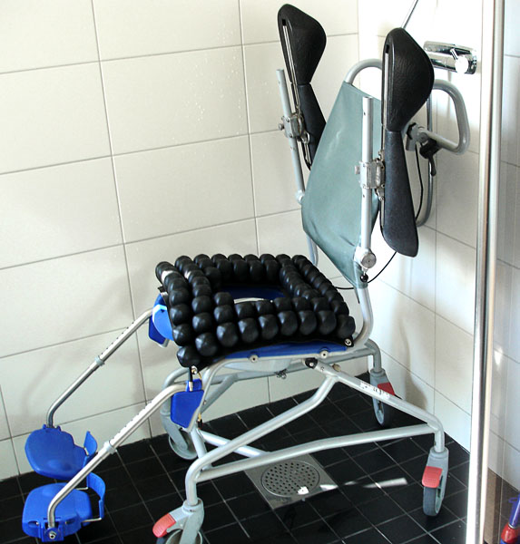 Dusch- och hygienstol med fyra små låsbara hjul och hög rygg av tyg. Armstöden är uppfällda och har skålformade polstrade underarm- och handstöd. På sitsen finns en Roho-dyna med hygienuttag.