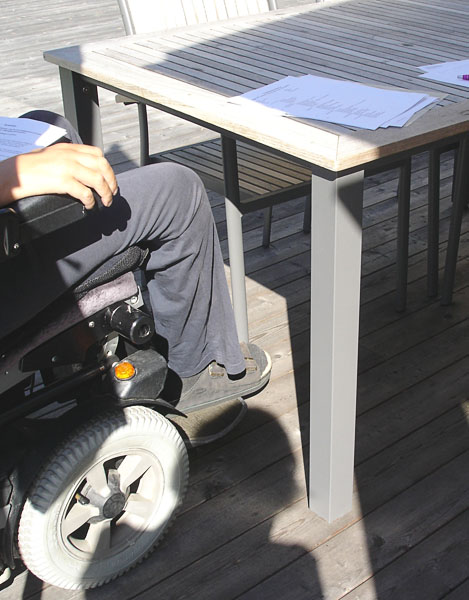 Användaren  sittande i elektrisk rullstol vid trädgårdsbord. Han kommer in under bordet med sina knä.