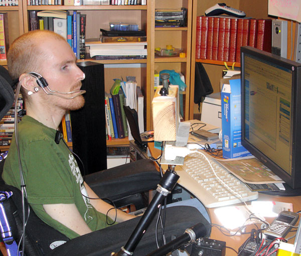 The user sits in front of the computer, wearing a reflector on his nose and a  headset for sip/puff control on his ear.