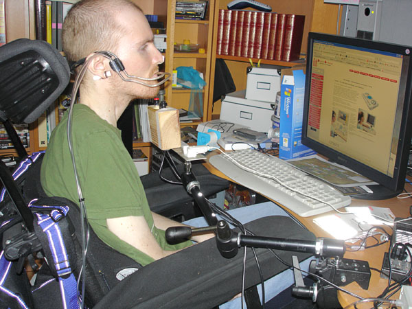 User sitting in electric wheelchair in front of computer. He has two suction and blow switches in his mouth attached to a headset. Under his chin he has a joystick and a microphone mounted on a table stand.
