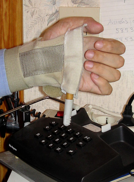 User with wrist bandage with attached universal holder. A small stick is attached to the universal holder so that it stands out on the ulnar side of the hand. The user dials a number with the telephone stick.