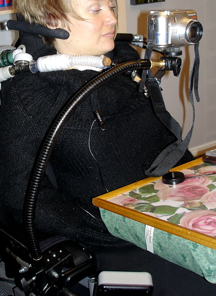 The user in the wheelchair, a flexible arm with a camera on it is screwed to the wheelchair's side support. The camera sits in front of the user's face.