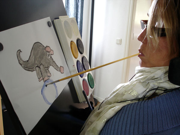 På en vinklad skiva sitter en teckning fastsatt med magneter och en målarlåda, användaren sitter framför den, hon har en pensel i munnen och ritar på teckningen.