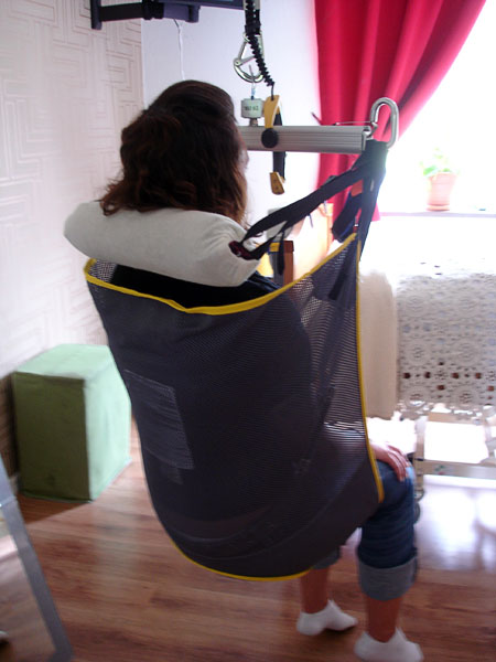 A person sitting in a ceiling lift The sling with the shoulders. Behind the neck there is a padded soft roller which is attached with straps to the sling bar.