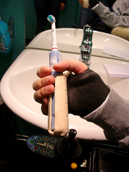 The toothbrush is in the users palm of the hand, with the holder, a hook, around the back of the hand.