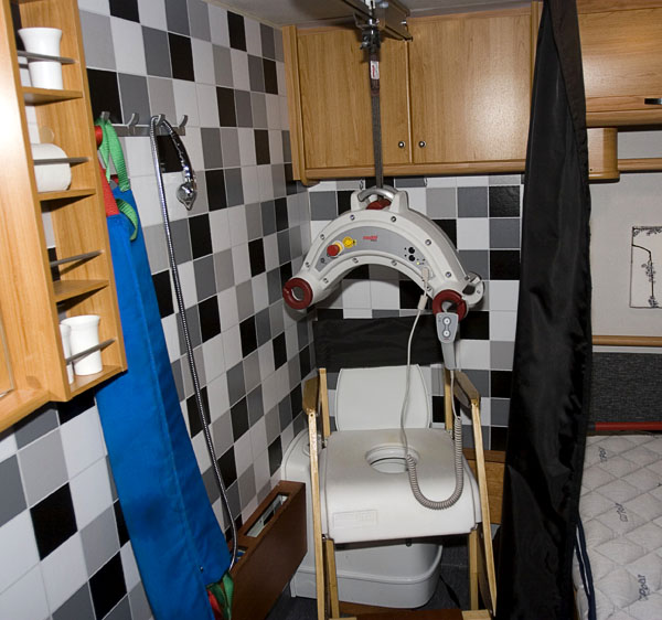 Shower area in RV with ceiling lift and shower seat with soft back, wooden armrest and padded seat with opening on toilet seat.