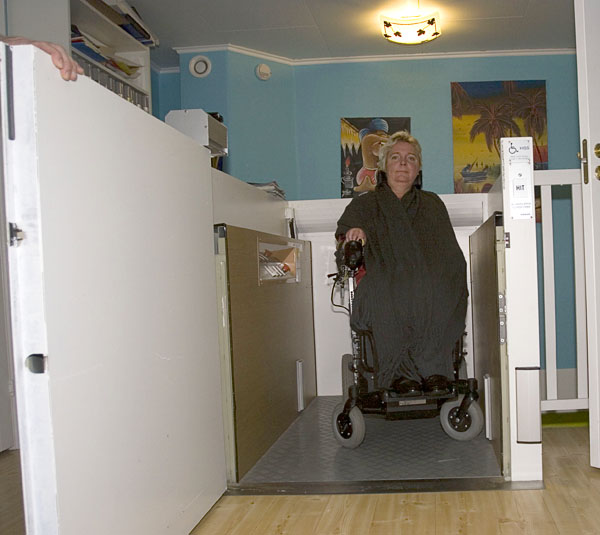 Helena in elevator; she presses elevator button with wheelchair frontwheel