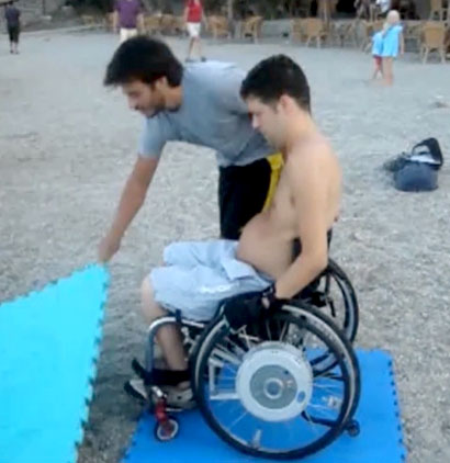 Användaren står med sin rullstol på en skumgummimatta. En hjälpperson lägger en annan skumgummimatta (som var bakom rullstolen) framför rullstolen.